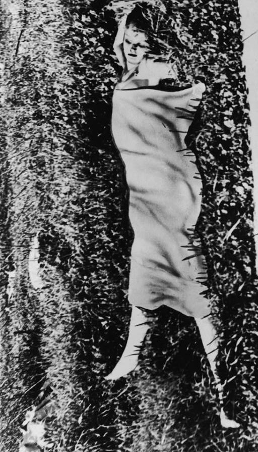 A historical black and white photograph of a person wrapped in a blanket, lying on the ground surrounded by grass.