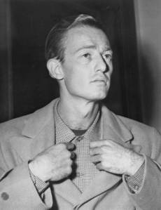 Black and white portrait of a man adjusting his shirt collar, looking thoughtfully to the side.