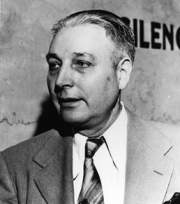 Black and white portrait of a man with short hair, wearing a suit and tie, appearing thoughtful or engaged in conversation.