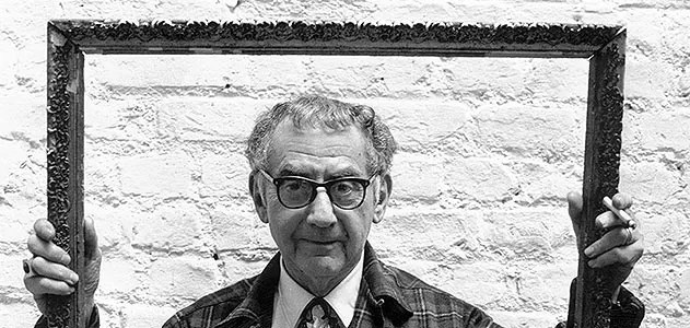 A man in glasses holding an ornate picture frame against a textured white wall.